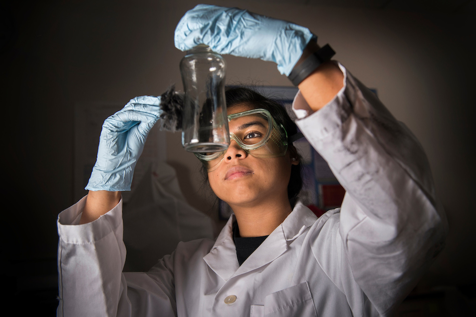 A student examines chemicals through a beaker while wearing a lab coat and goggles. 