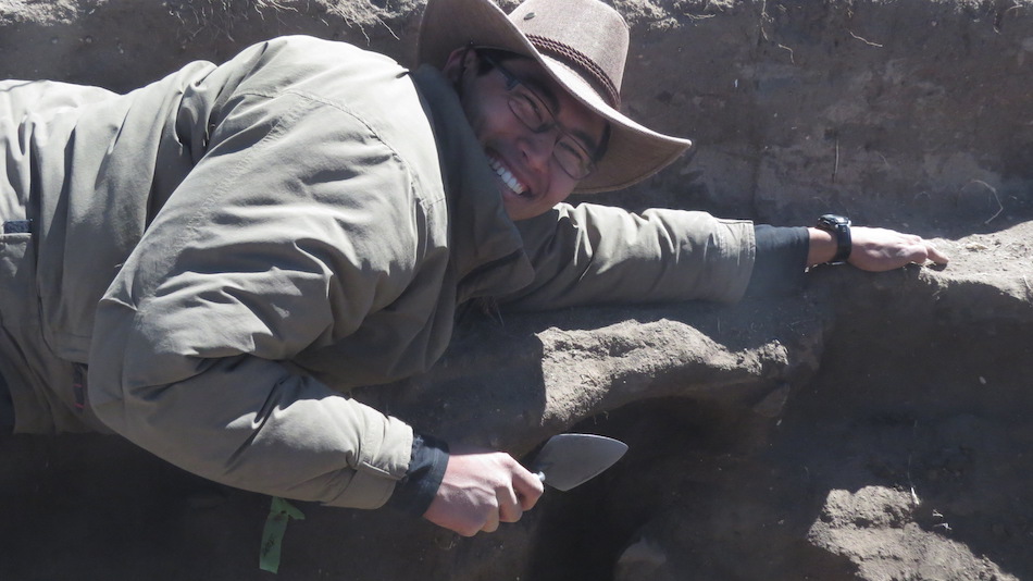 A student in a sunhat leans over a sandy trench with a hoe, smiling. 