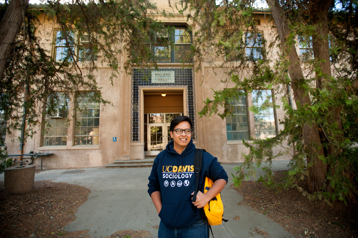 A sociology grad student wearing glasses and a sociology sweatshirt stands in front of a campus building with a backpack.