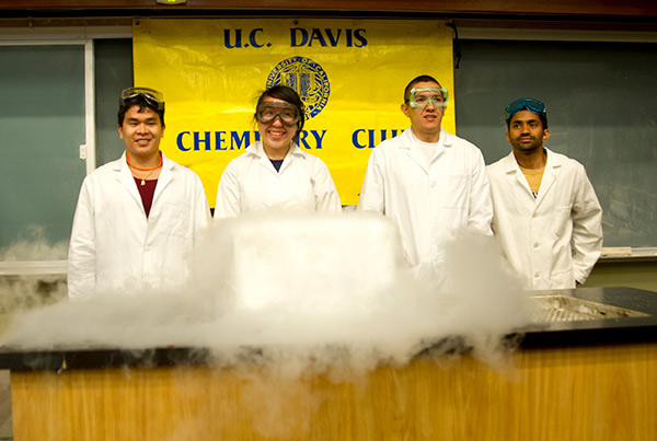 Four people in white coats from the UC Davis Chemistry Club standing behind cloud-producing chemicals on a table