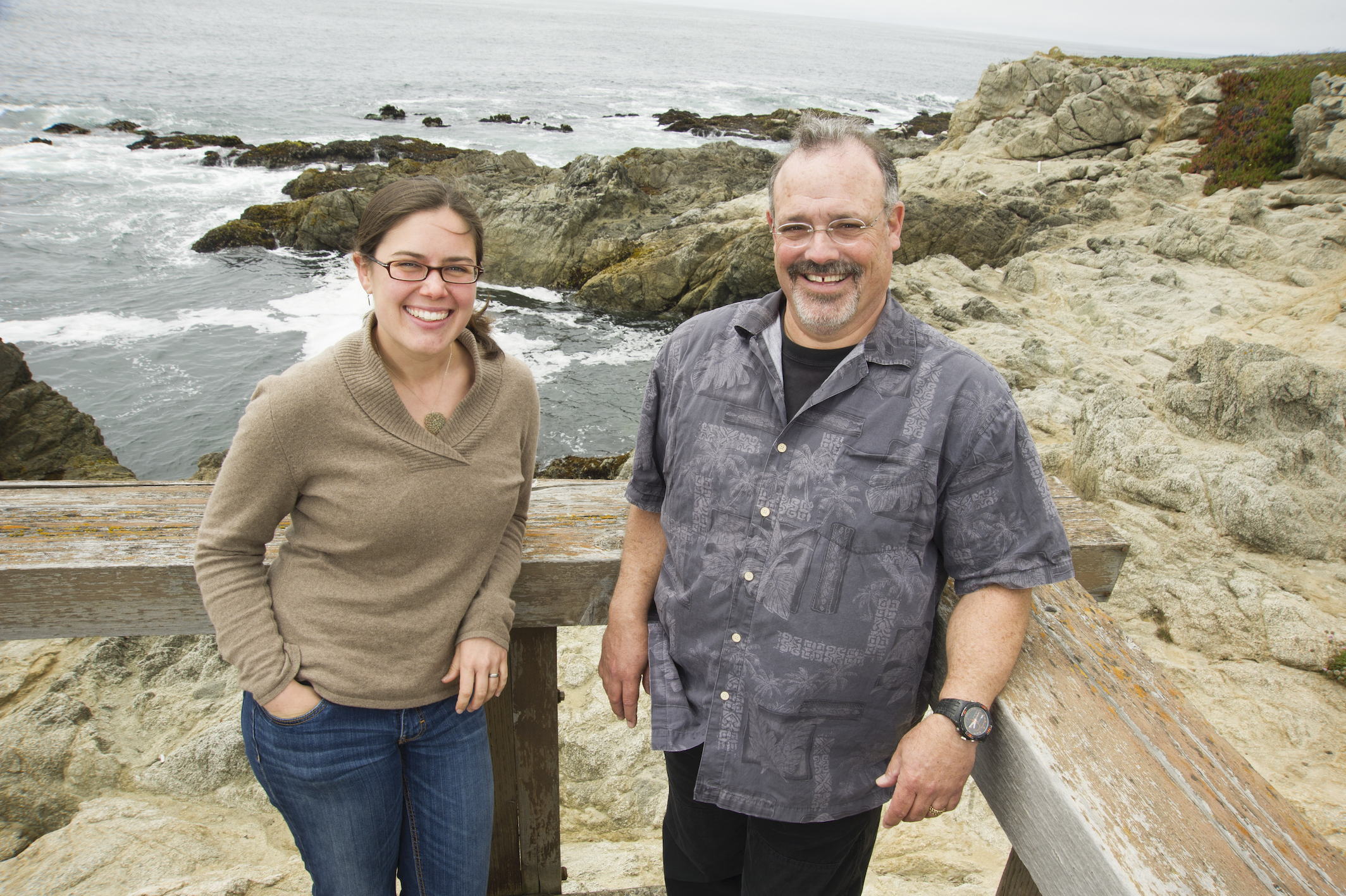 Female and male scientists face camera smiling on a deck with ocean in background
