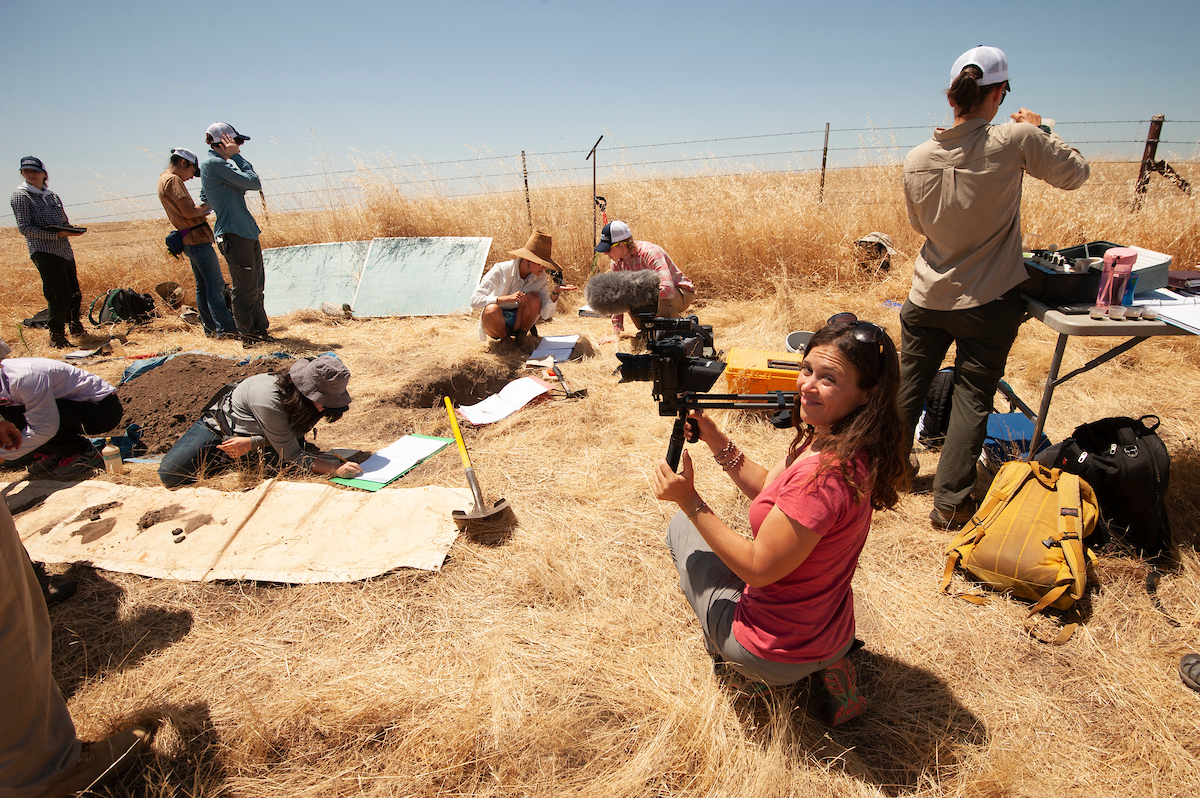 Woman in red shirt with video camera smiles while soil science students sample soil in dry straw landscape