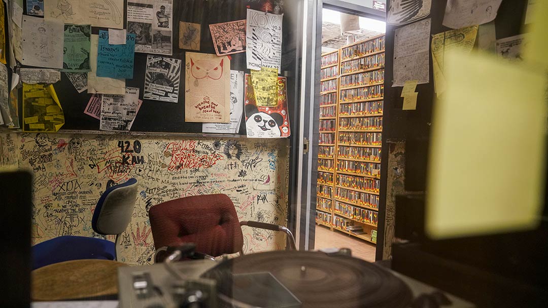 Photo from inside a listening room at the radio station. A turntable is seen in the foreground, with an old swivel chair behind it. The wall behind has handwritten messages scrawled on every inch of space, as well as bulletin boards with flyers. An open door reveals a hallway with floor to ceiling racks of cassette tapes lining the walls.