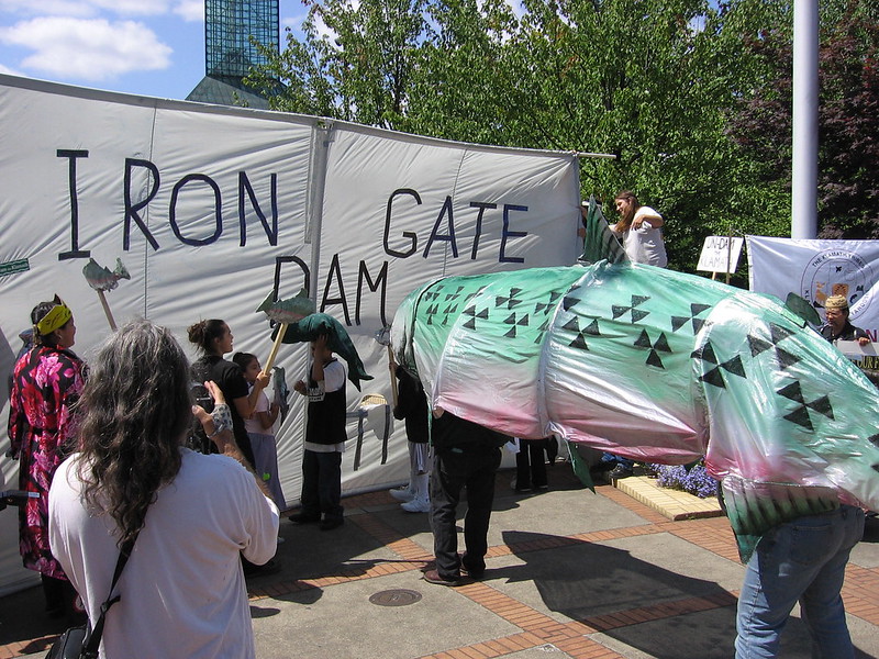 Klamath Basin tribes protest in 2006 with banner representing Iron Gate dam and large salmon costume appearing to be blocked behind it