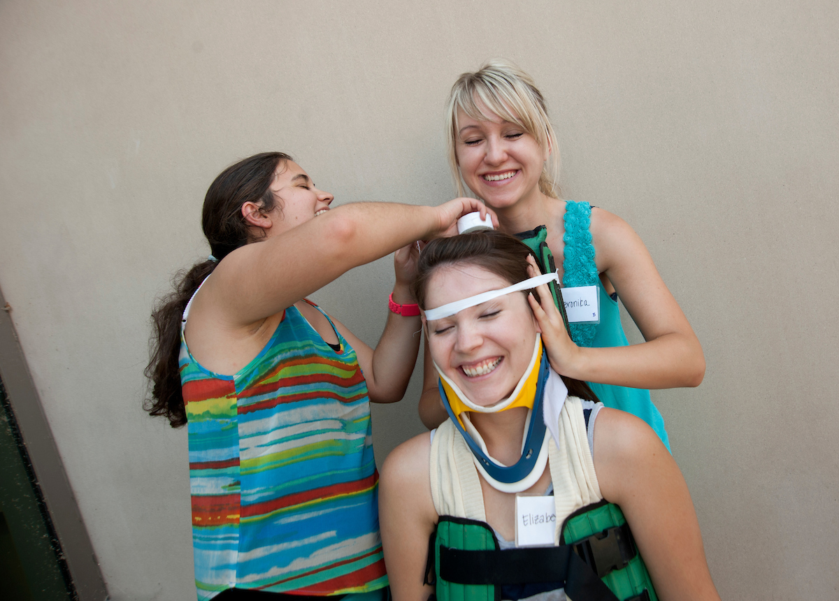 Two students laugh as they practice bandaging the head of another student.