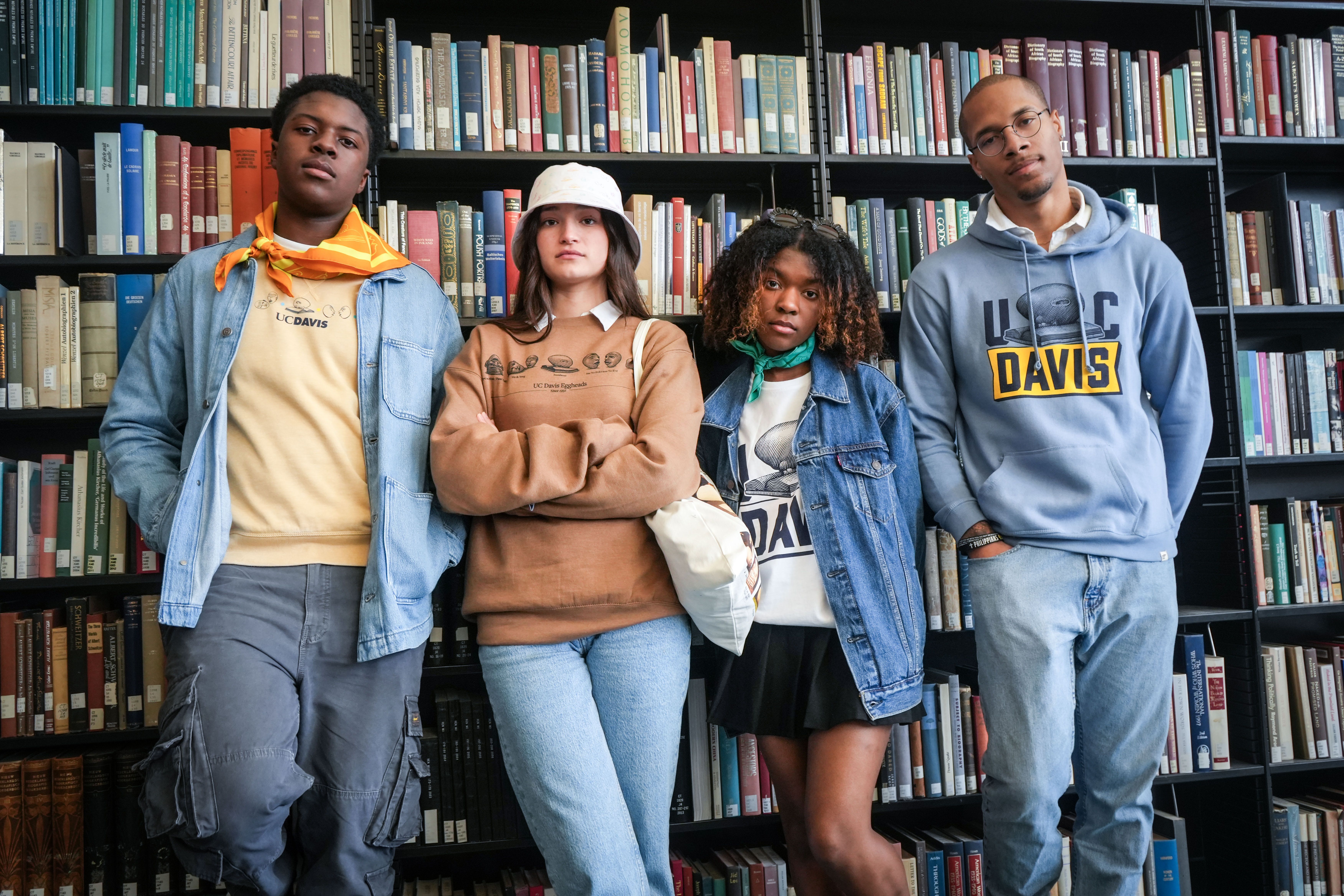 four students stand next to each other and face the camera in a library setting. all four are wearing apparel from the new Arneson Egghead Collection featuring the Egghead sculptures made by former UC Davis faculty Robert Arneson.