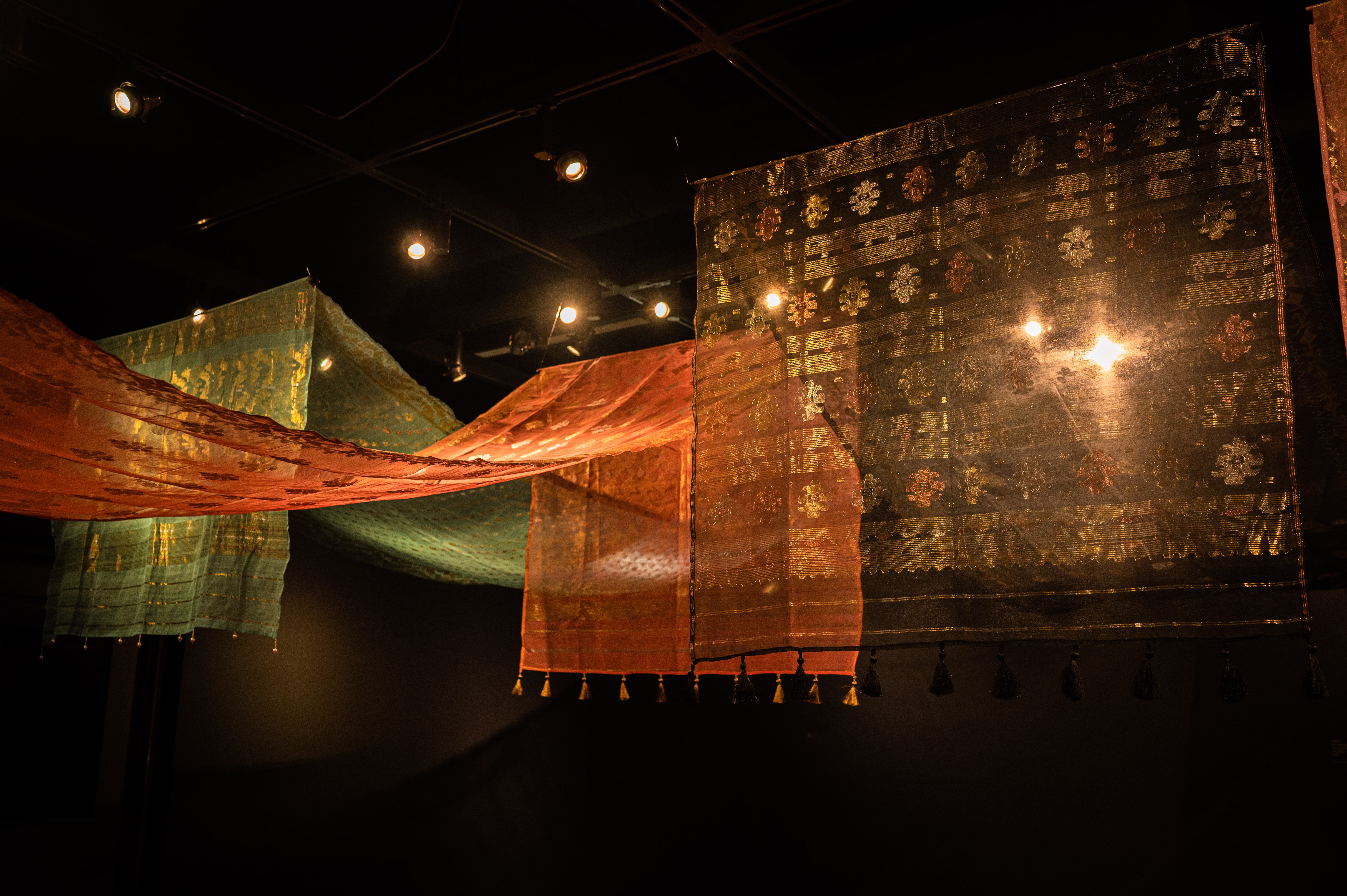 Three Jamdanis, blue, pink, and black are strung along the ceiling against a black background. Each fabric contains gold patterns and light shines through the black Jamdani.