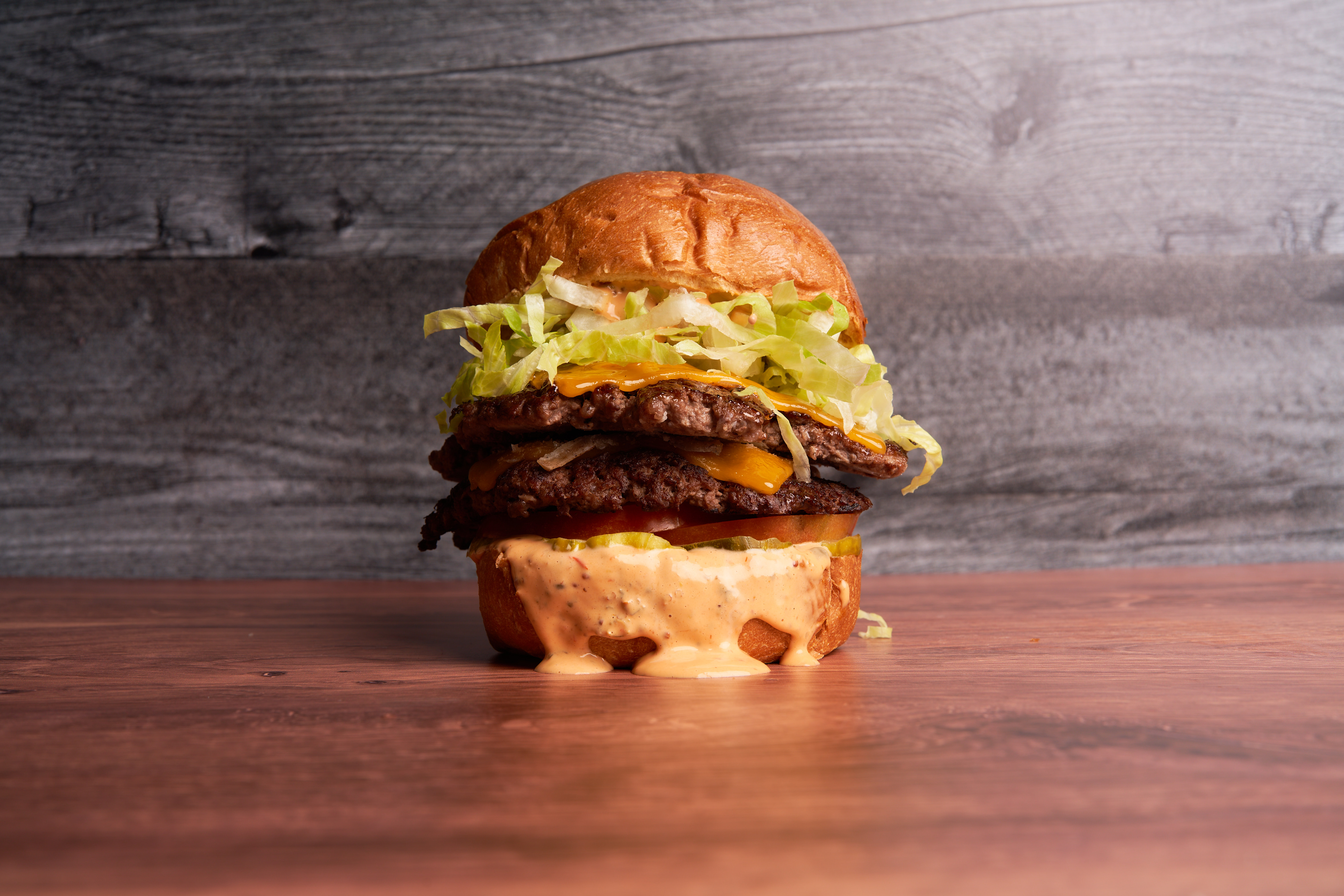 A well-stacked smash burger sits centered on a wooden table, served with numerous toppings and ready to eat.