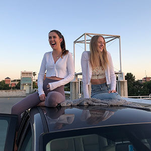 Two female students sit on the hood of a car around sunset, laughing.