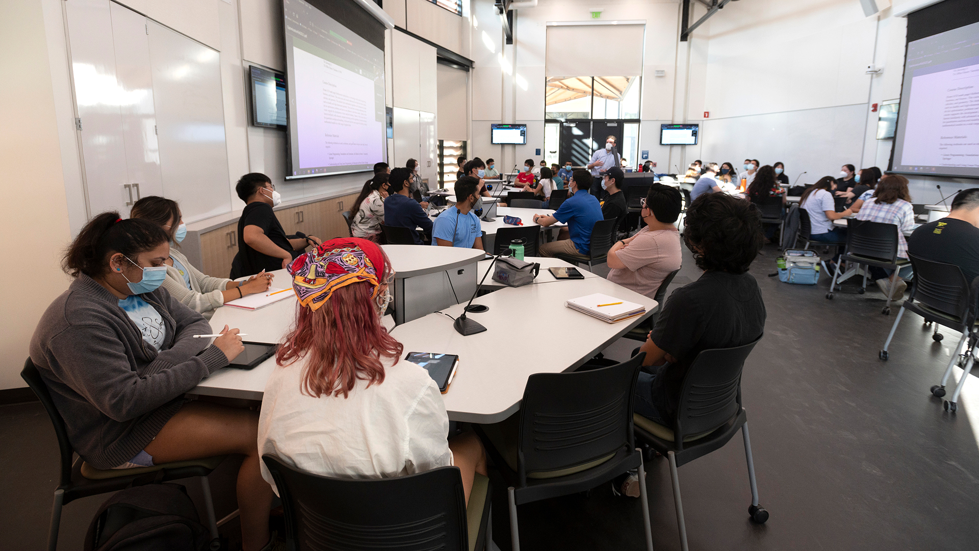 Photo inside a classroom during a class. Students are seated together around tables, interacting. Tables have microphones for each student and areas to plug in laptops. A lecturer is walking and talking in the background.