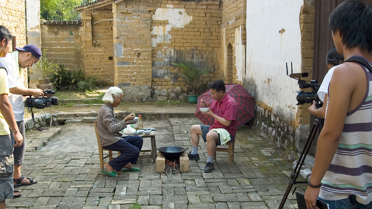 Martin Yan and a woman sit as they eat a meal outside in a Chinese village 