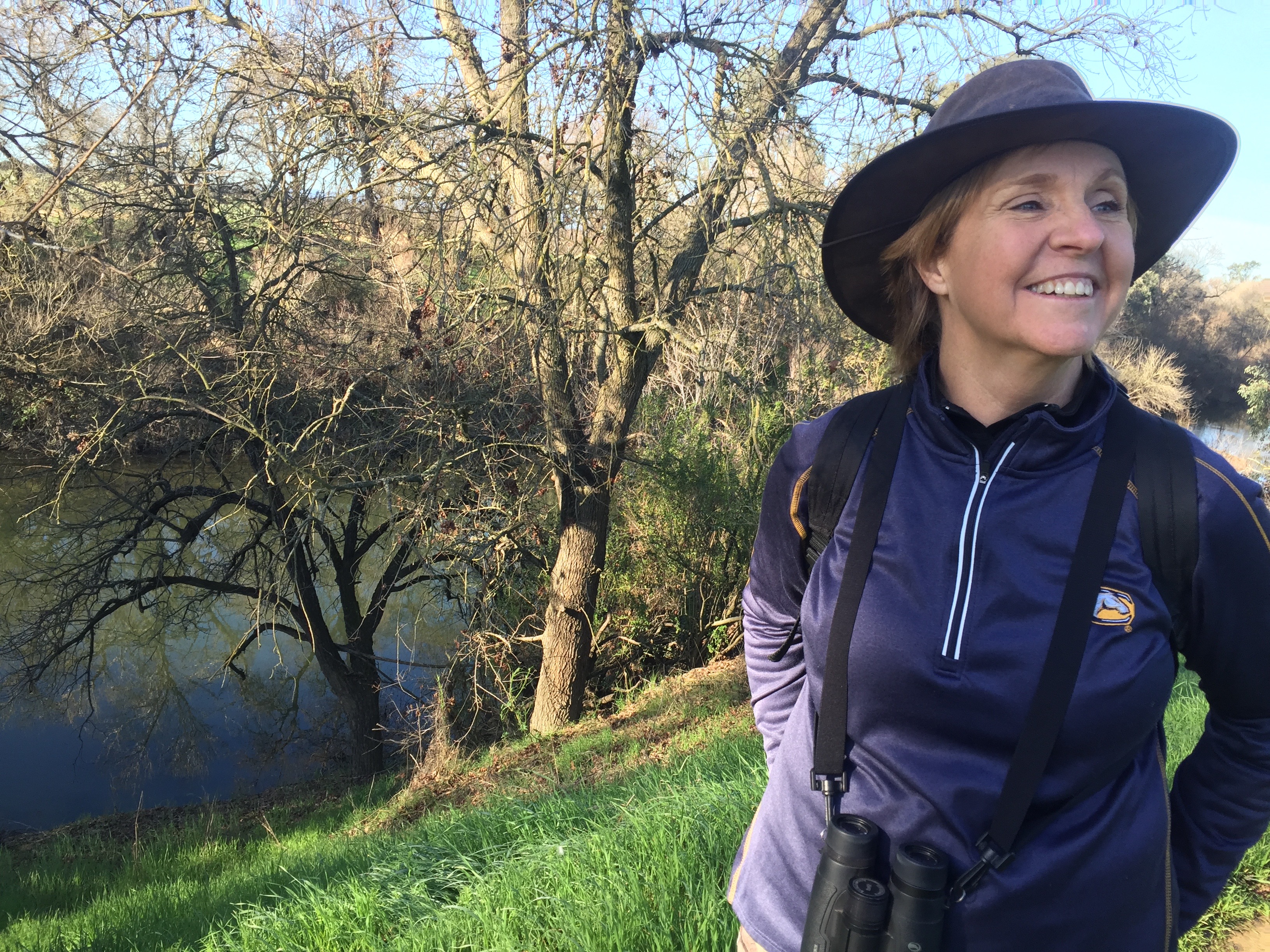 woman wearing hat smiles standing along side a wooded creek