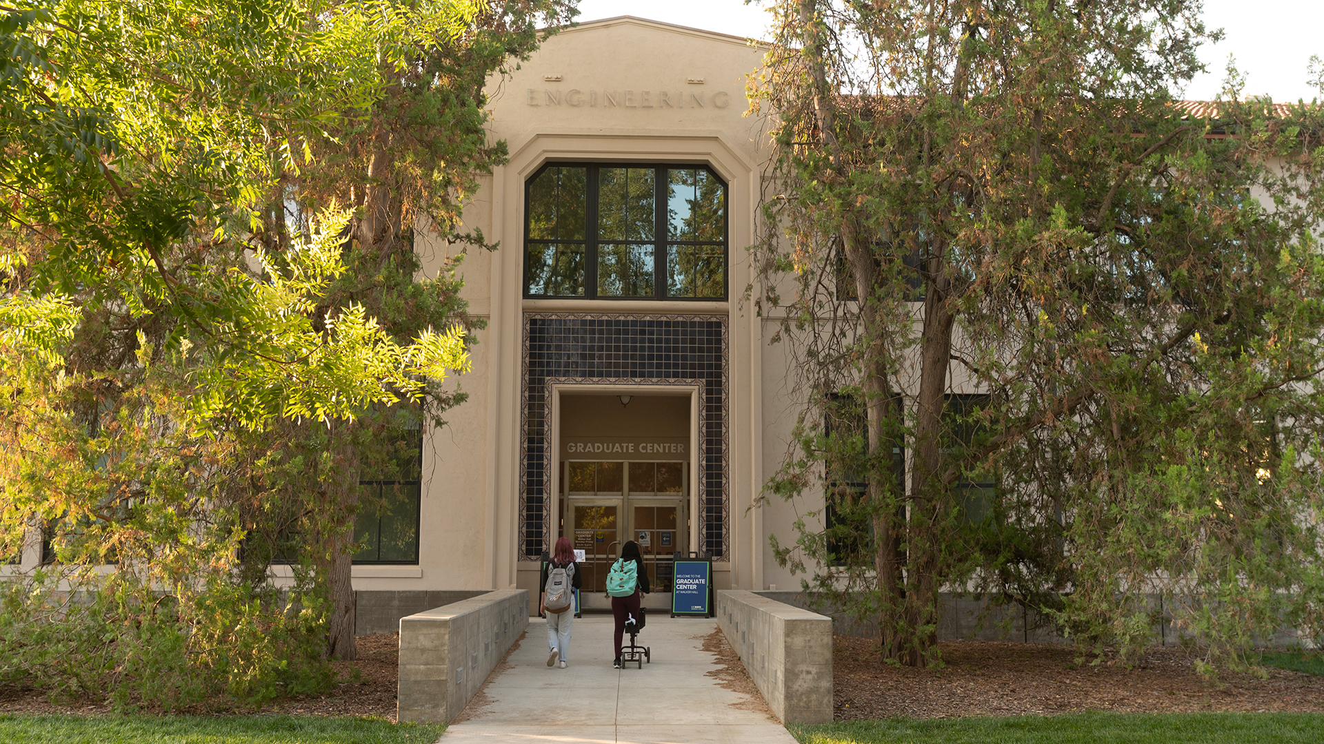 Photo of an older building facade with the word Engineering in old letters high above an entrance where double doors are framed by bright blue tiles. A path leads directly to the doors through a short lawn. Large oak trees flank the entrance with golden light. In a modern typeface, it says Graduate Center above the doors.