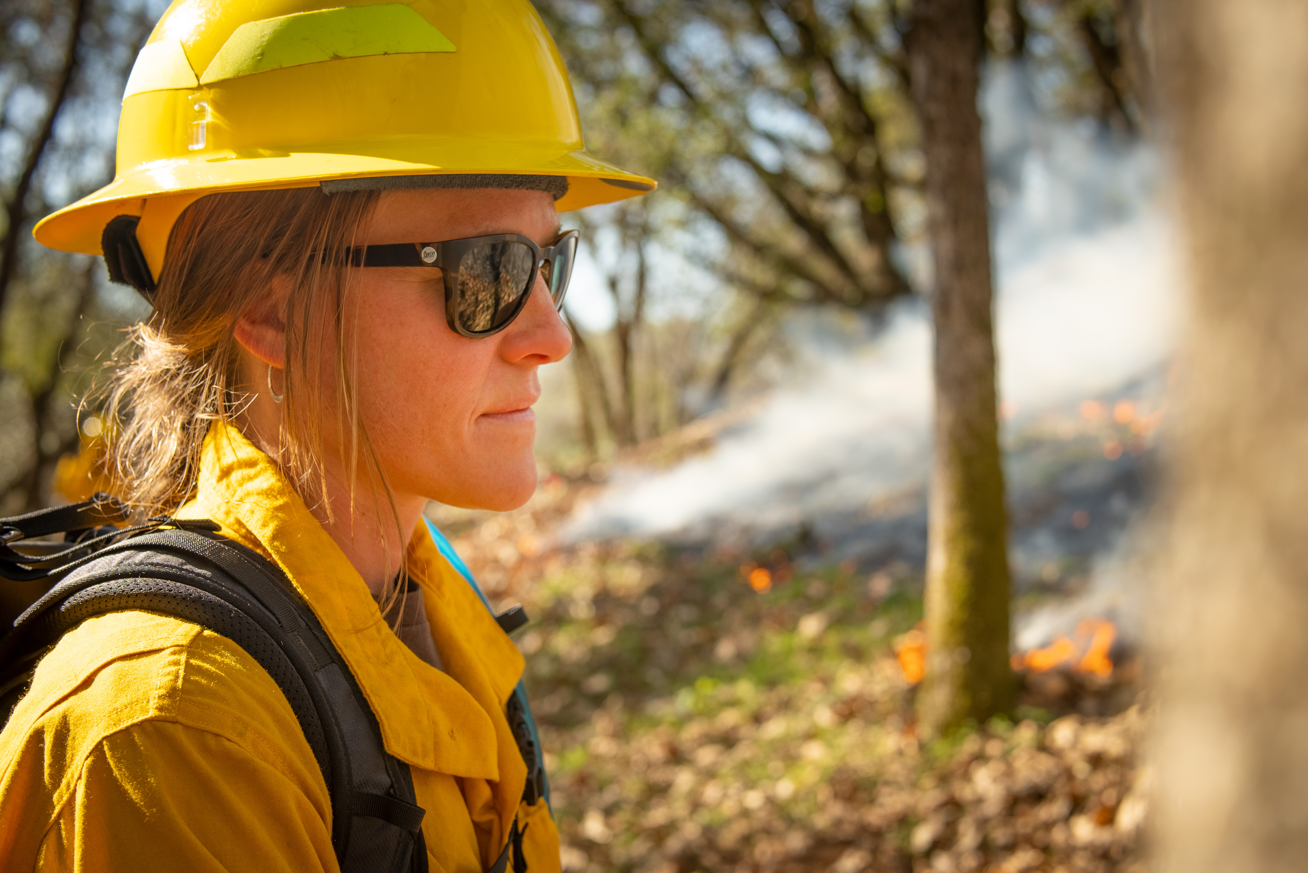 Woman in yellow hard hat and yellow shirt walks in forest with smoky background during prescribed fire