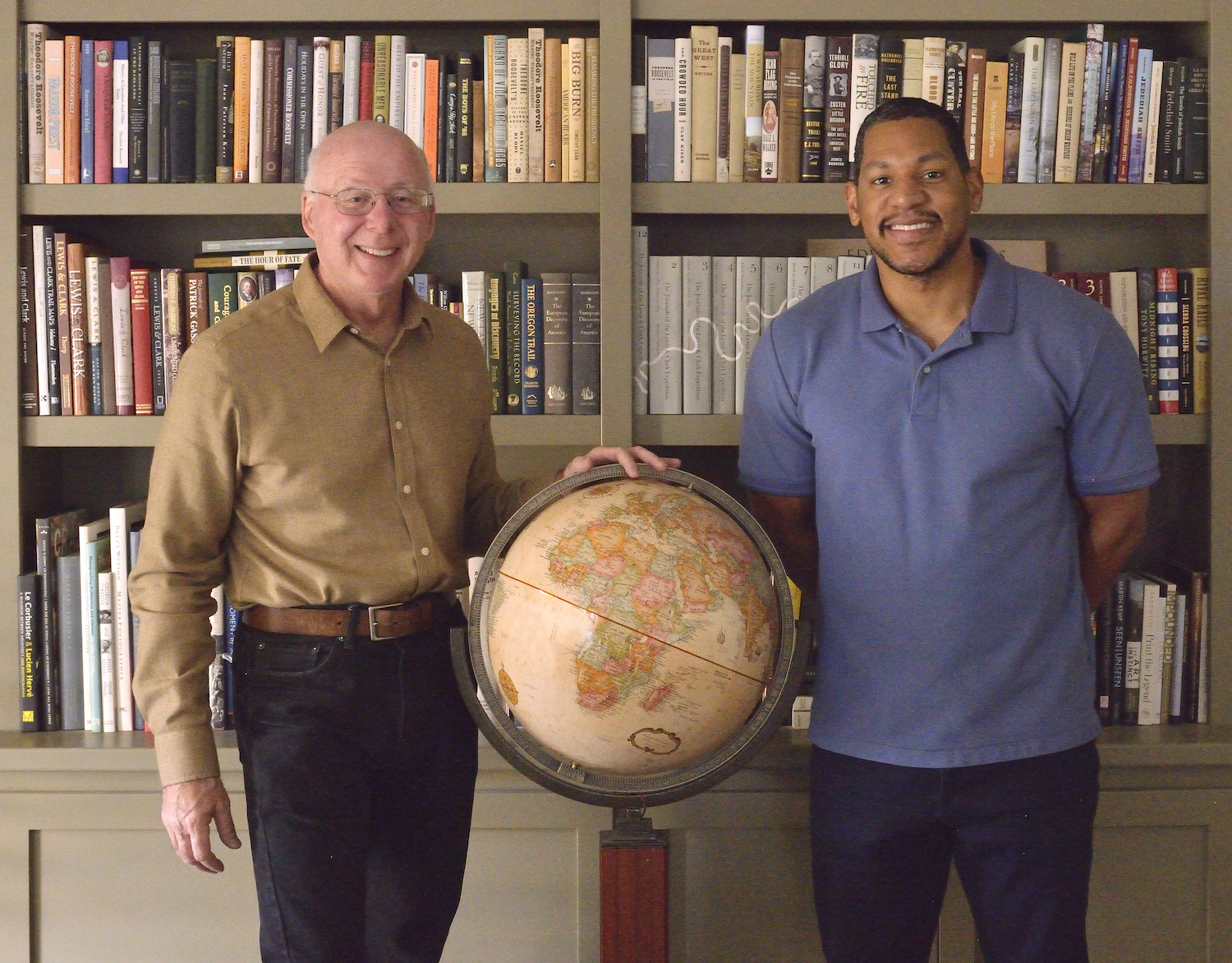 From left, white male professor in beige shirt and black pants stands alongside black male Ph.D. student in blue shirt and blue plants, a globe between then and shelves of books in background. 