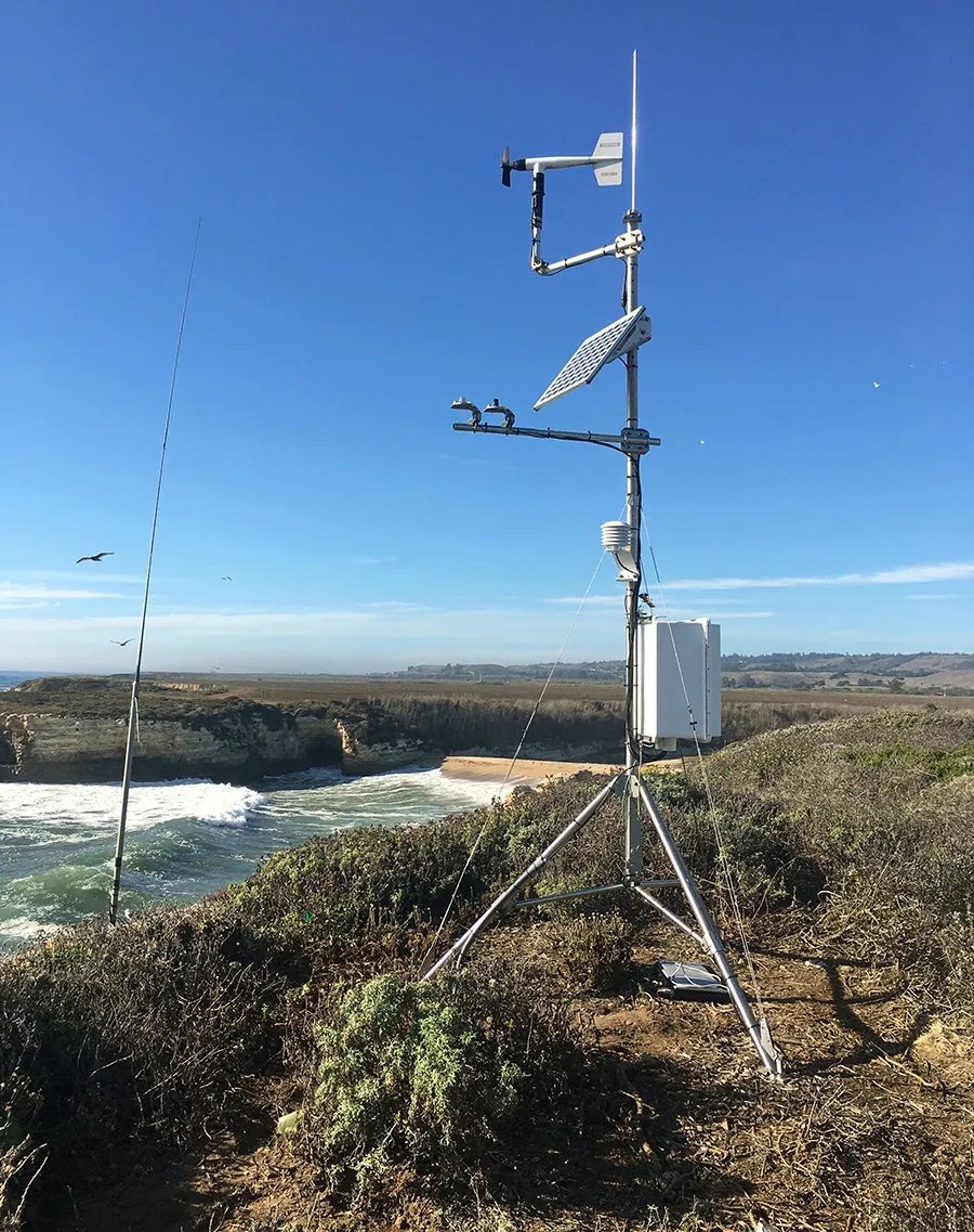 A climate station consisting of a tripod, solar panel and other instrumentation overlooks shrubby coastline and rolling waves at Younger Lagoon Reserve