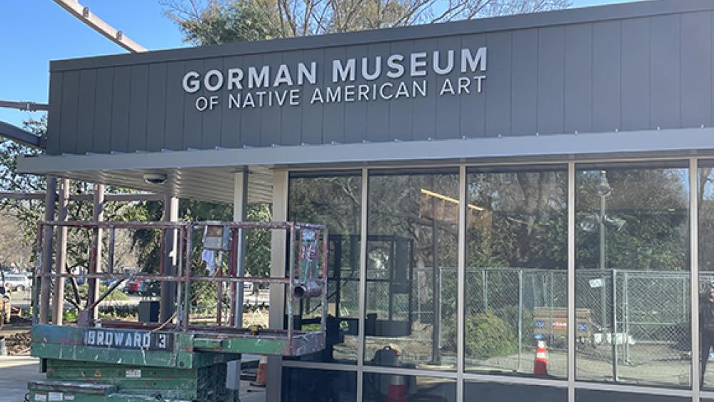 One side of the museum. Vertical windows make up the walls and a gray trim along the top has a sign that reads "Gorman Museum of Native American Art." Construction equipment lies nearby.