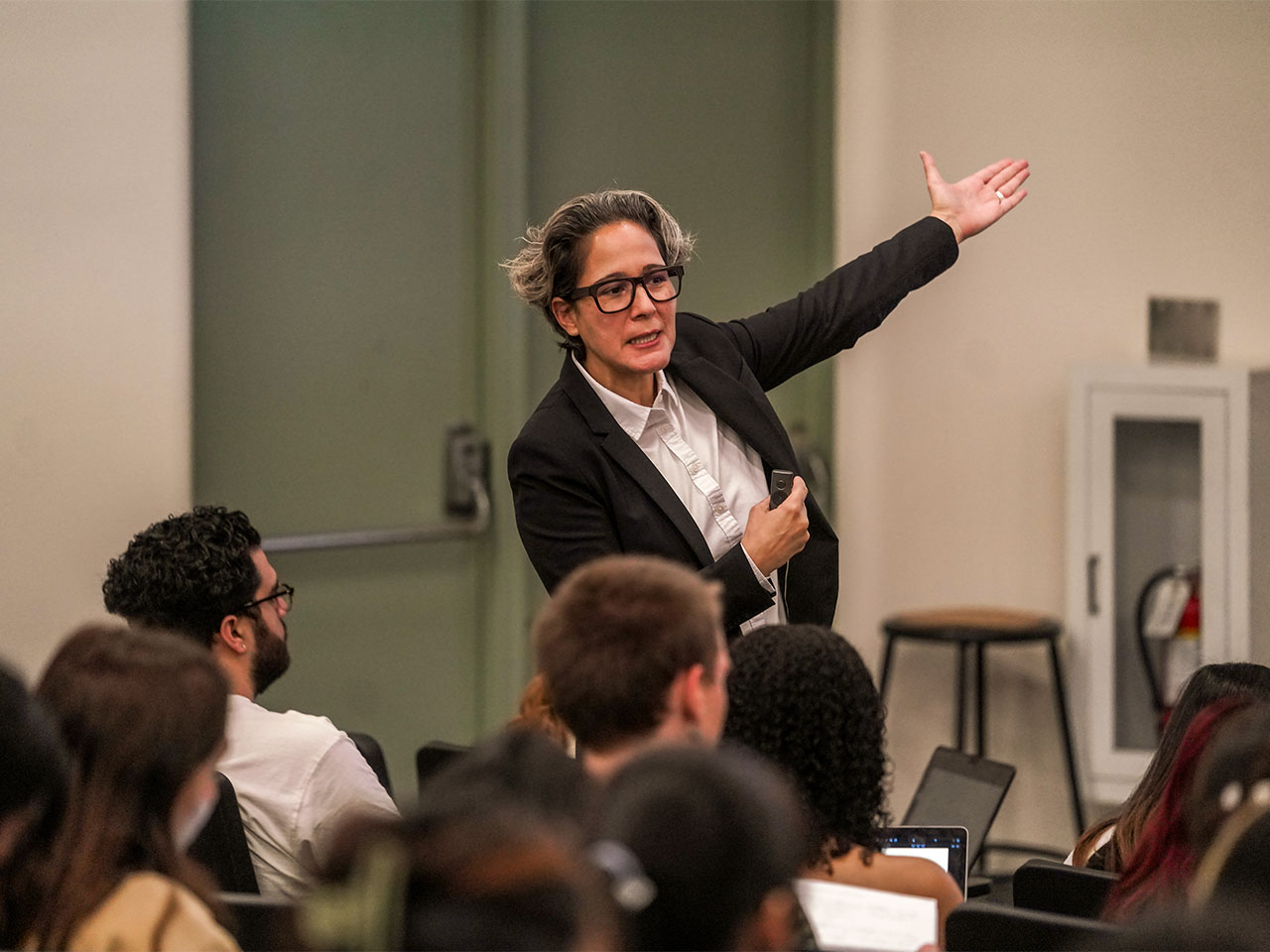 A professor with short gray hair, wearing square-rimmed glasses and a black blazer points to a presentation slide off-camera while teaching an art history class at UC Davis.