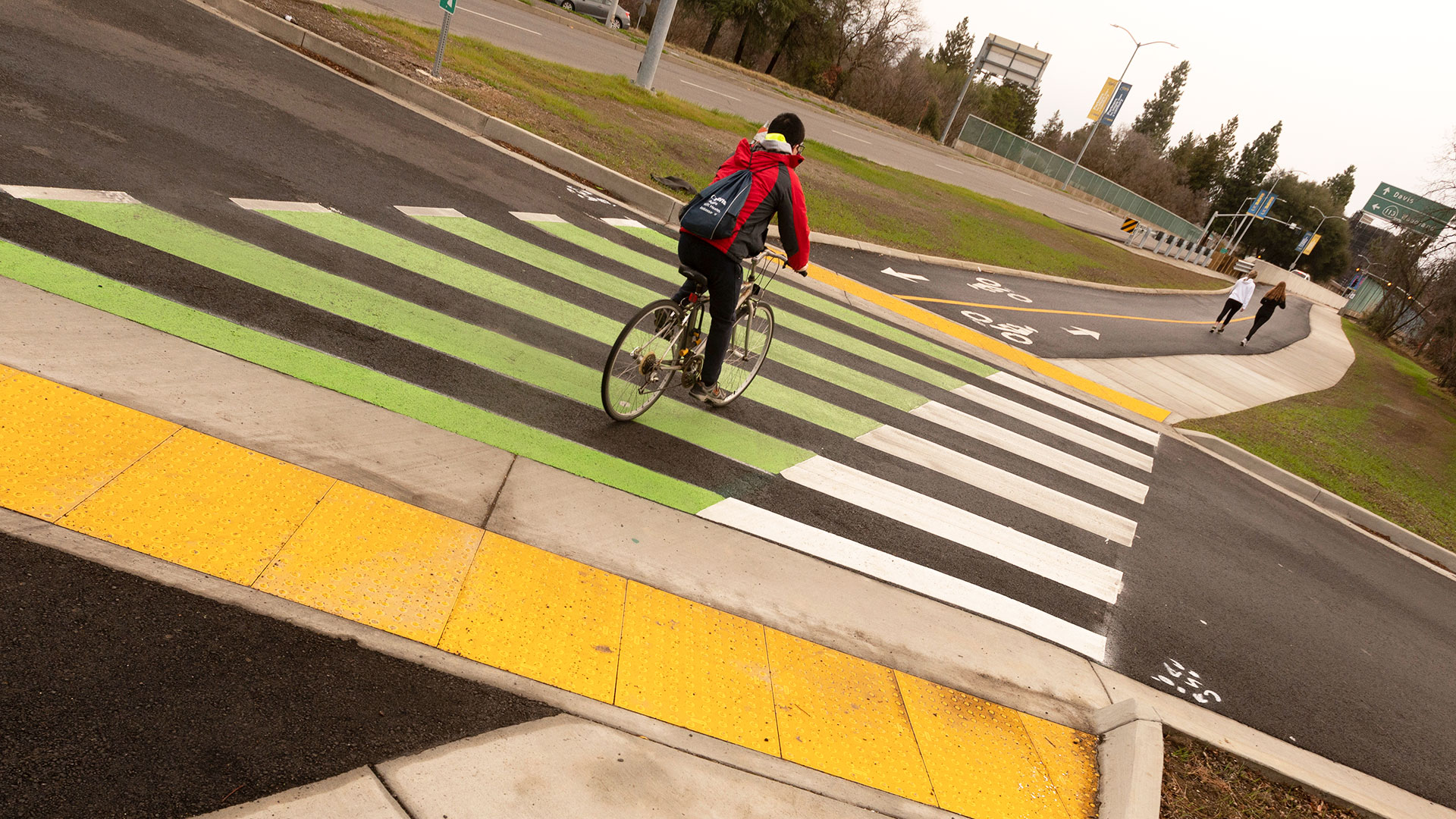 Photo of a bicyclist riding away from the camera on a bike path that intersects a street. The intersection has green and white striped paint across it and yellow bumped plastic at the edges.