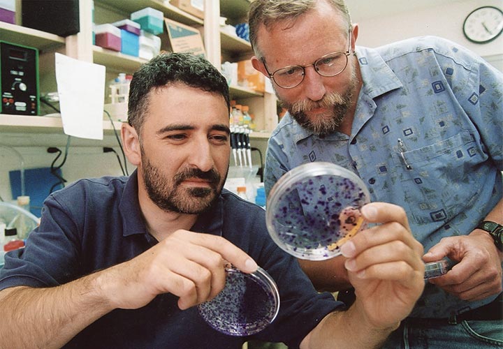 A man holds up a large magnifying glass and looks through it while another man stands behind him looking through the glass too.