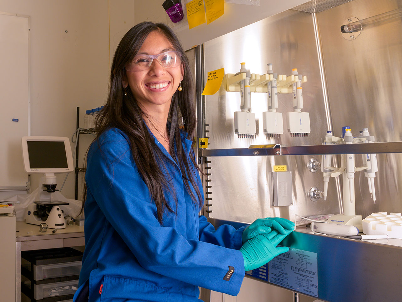 A smiling scientist in a blue lab coat working in a laboratory setting, with safety goggles on and handling lab equipment.