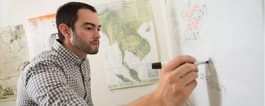 student writing on a dry erase board with pictures of maps in the background as part of the linguistics graduate study program