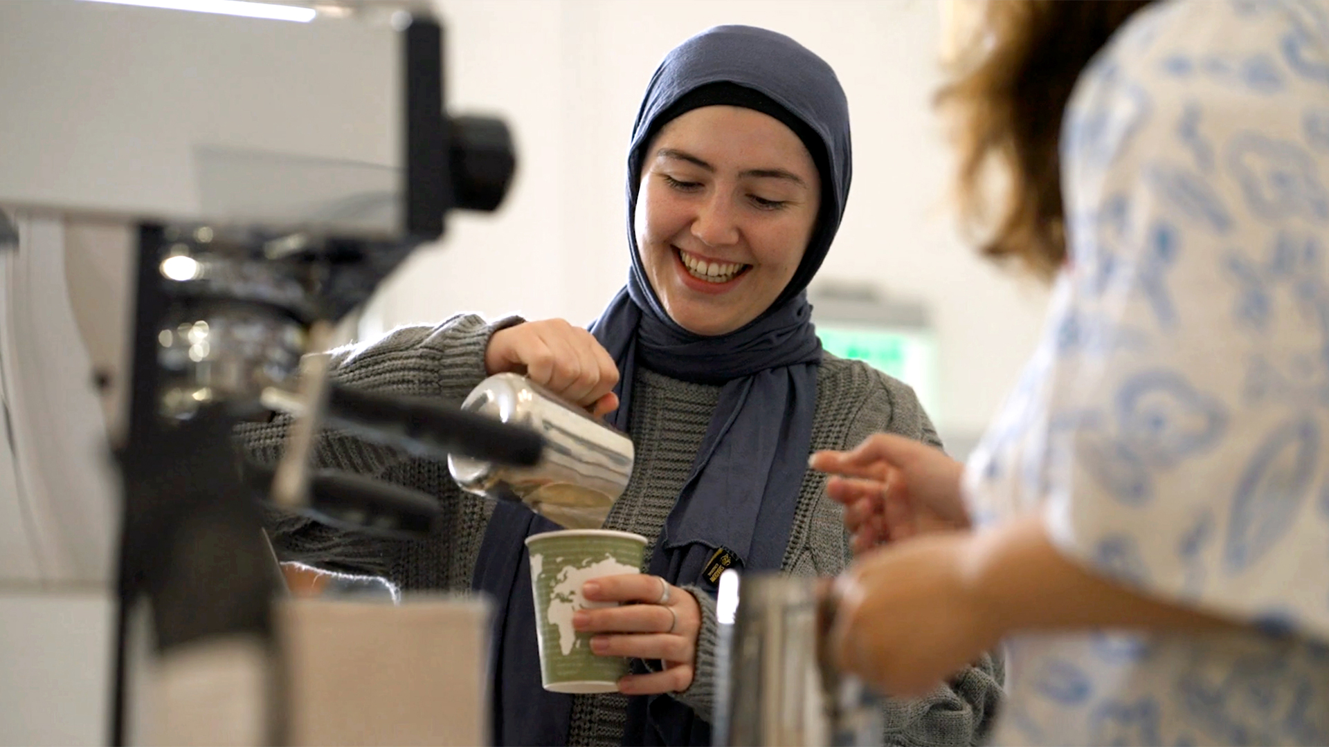 A smiling student in a hijab pours foam into paper cup with a green and white printed design of the Earth's continents.