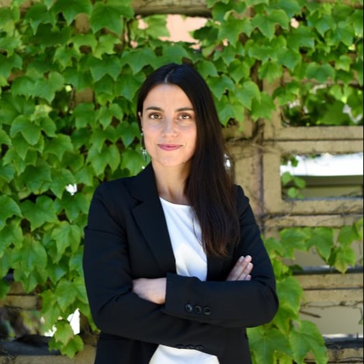 Head shot of Cristina Lazcano, soil scientist, in black jacket, white shirt, folded arms, with vines in background