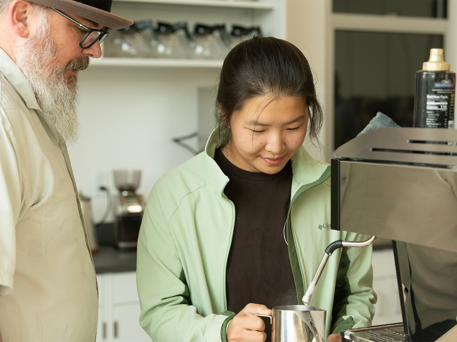 Timothy Styczynski, a man with a white beard, glasses, and light button-down shirt, watches as student Peyton Yang, wearing a black shirt and mint green jacket, uses an espresso machine to froth milk for a latte.