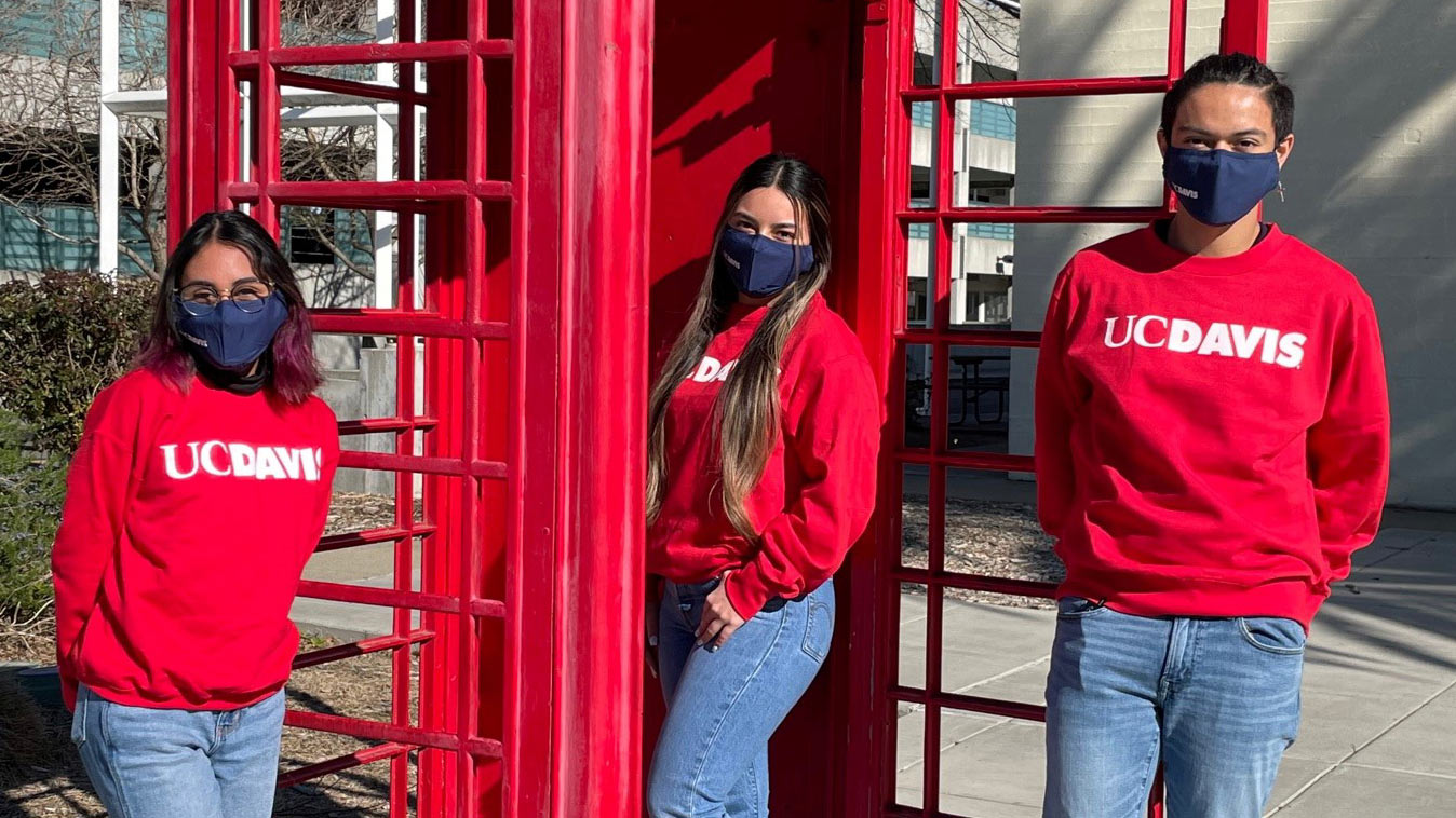 Two women and man in red "UC Davis" sweatshirts, posing at London phone booth