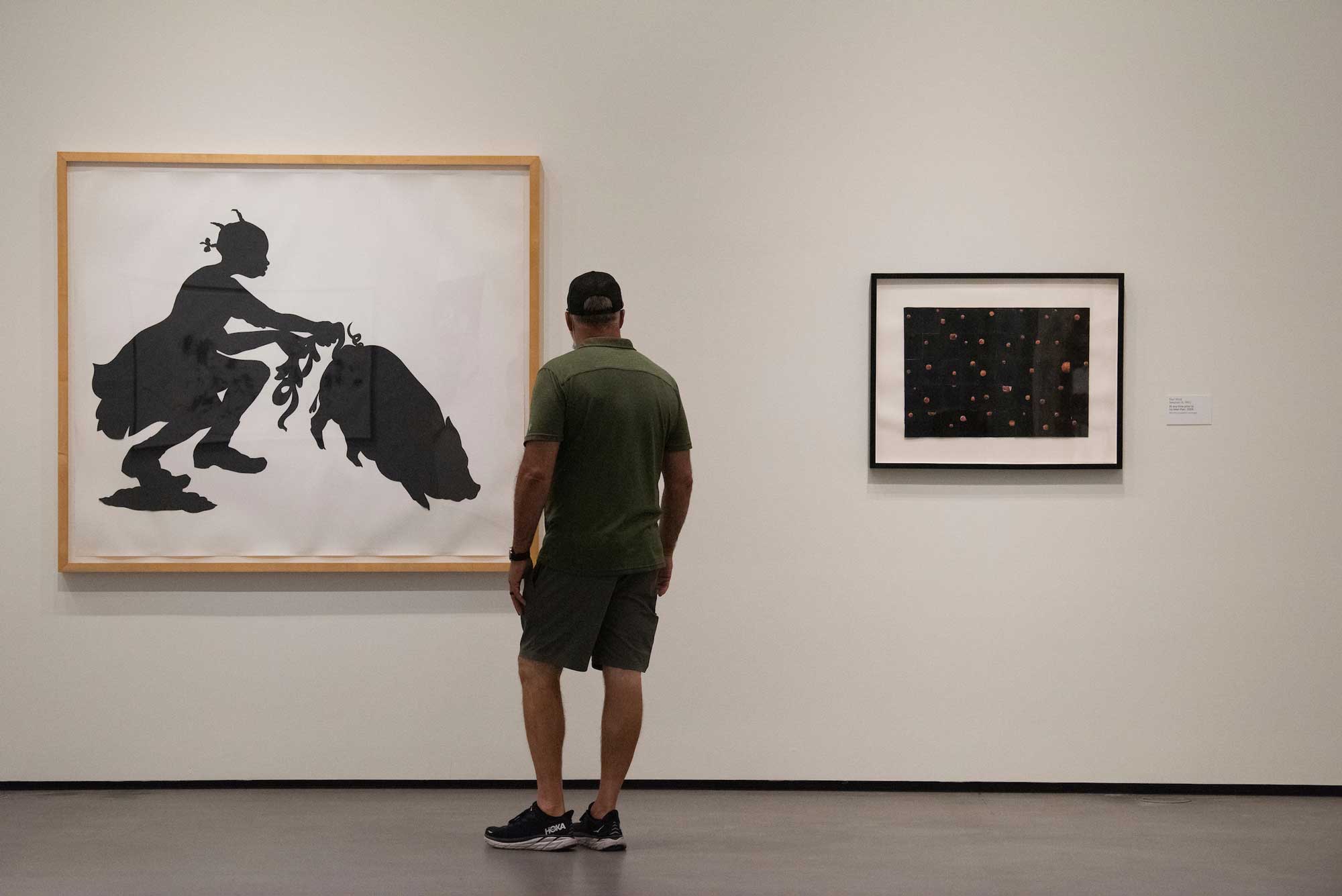 A man stands in front of Kara Walker's artwork of a black silhouette of a young girl playing with the entrails of a pig.