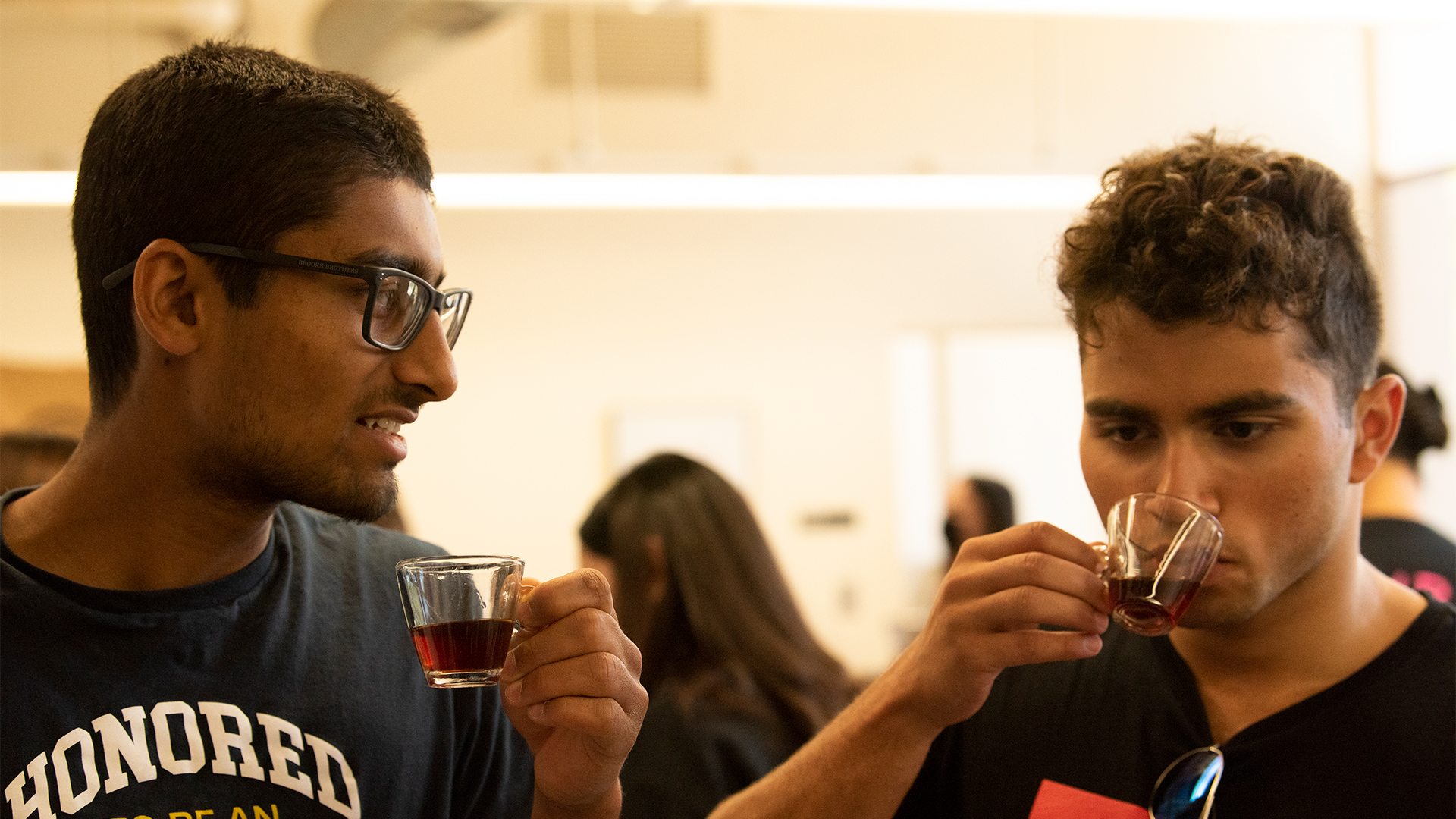 Two students concentrate as they taste coffee in small glasses.