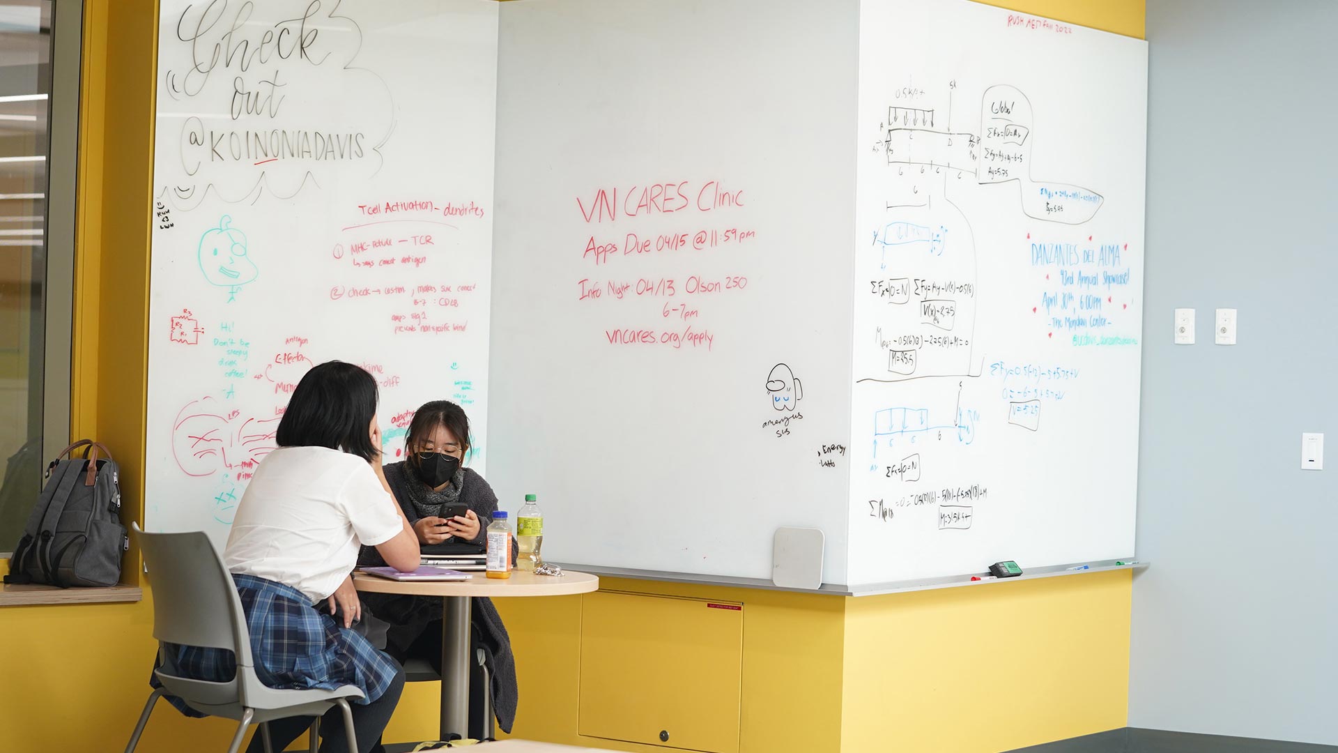 Two students sit at a cafe table in a student lounge area. A large wraparound white board on the wall has messages and equations scrawled on it.