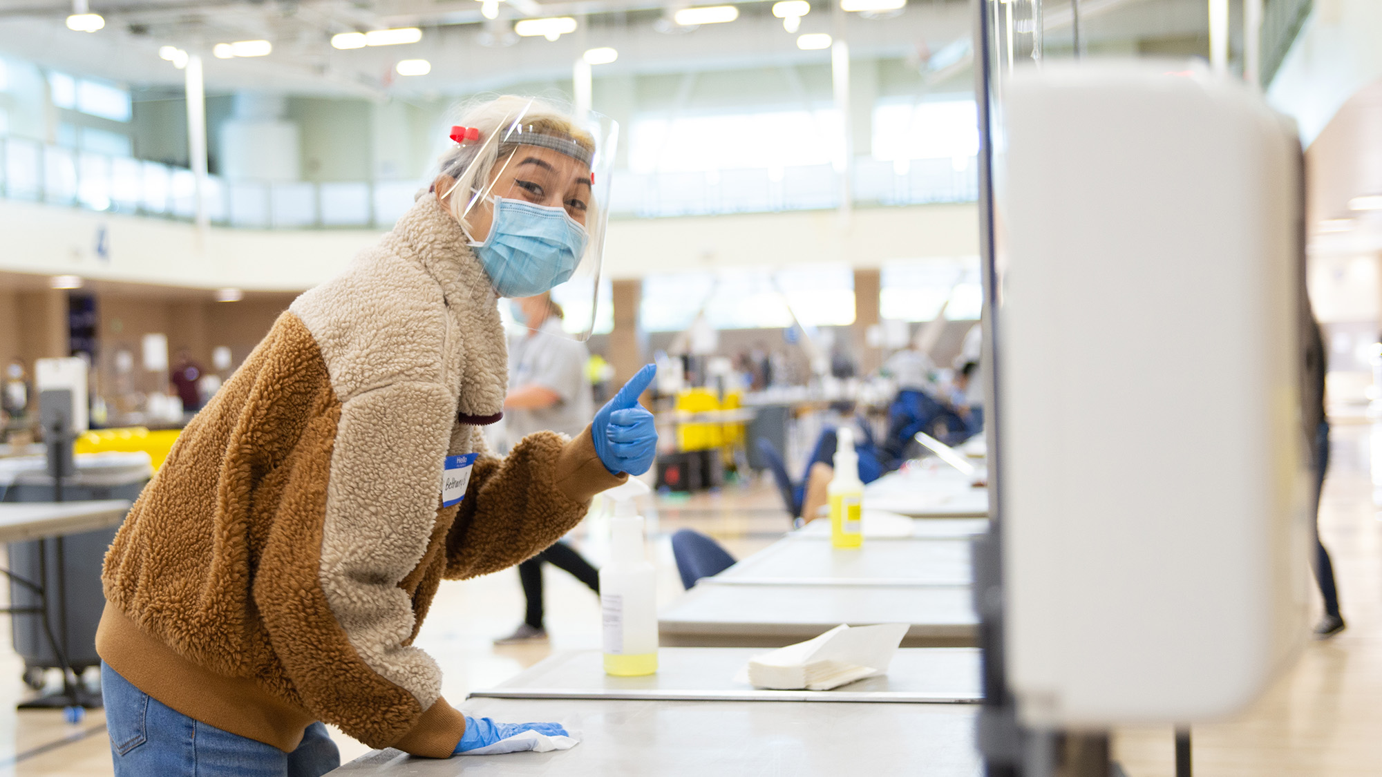 Worker in face mask cleans table while giving thumbs-up.