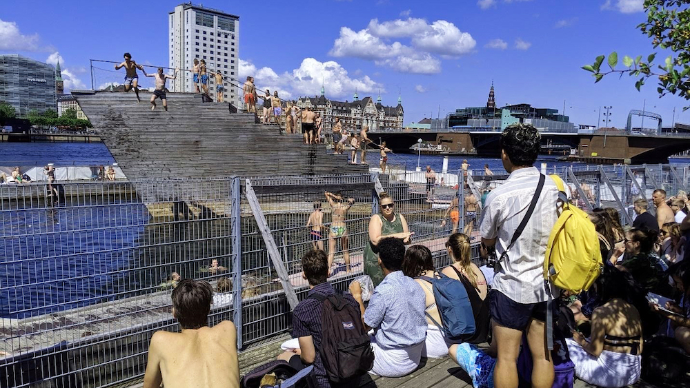 A crowd gathers around a river while a few people jump in. 