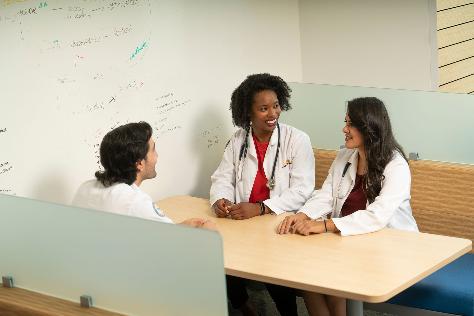 three med students sitting at a booth style table with a white board on the wall behind them