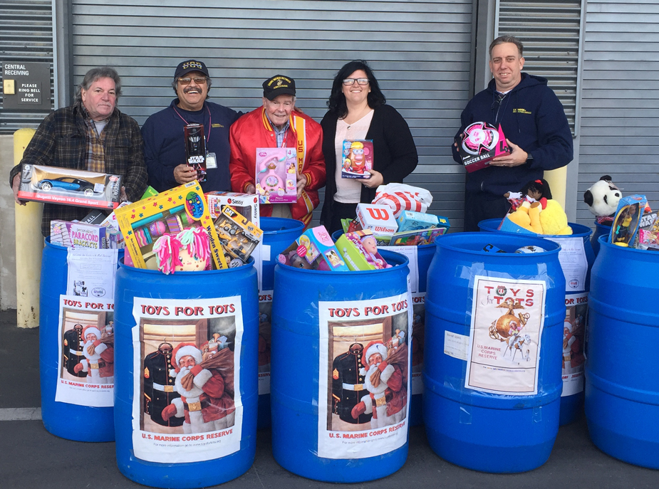 Five people stand behind Toys for Tots barrels filled with toys.