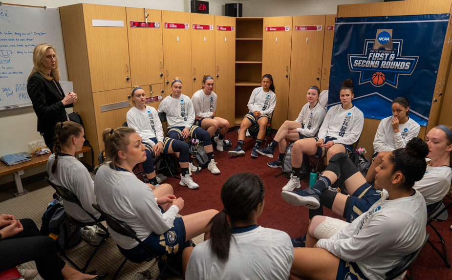 Head coach addresses team, seated in circle, in locker room.
