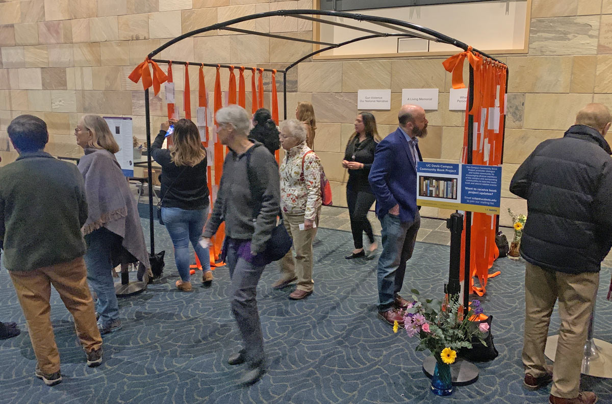  orange ribbons with tags attached, hanging from metal structure, with a dozen people viewing.