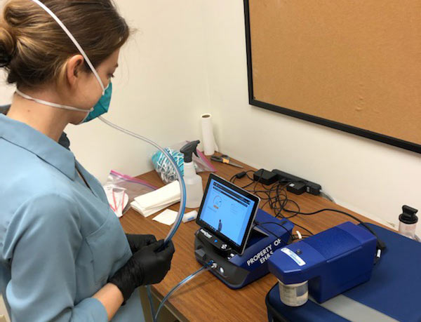 Woman wears N95 respirator that is connected via air tubing to a small electronic measuring device.