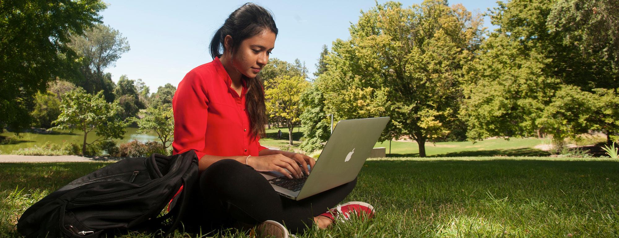 A student sits in the arboretum on a nice patch of grass surrounded by trees working on her laptop