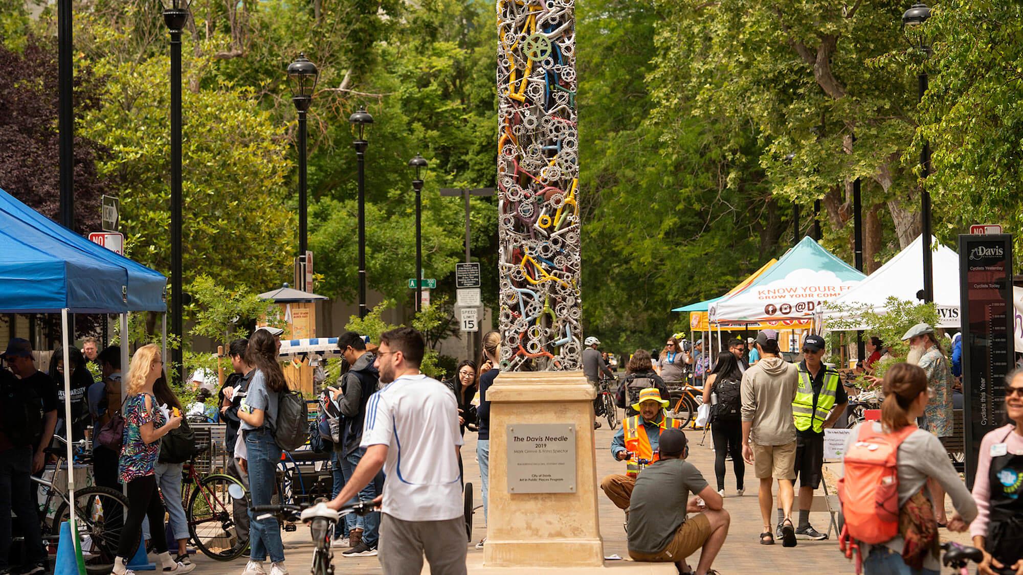 A scene of downtown Davis, Ca, full of students, locals and a unique obelisk shaped sculpture made of bicycle parts