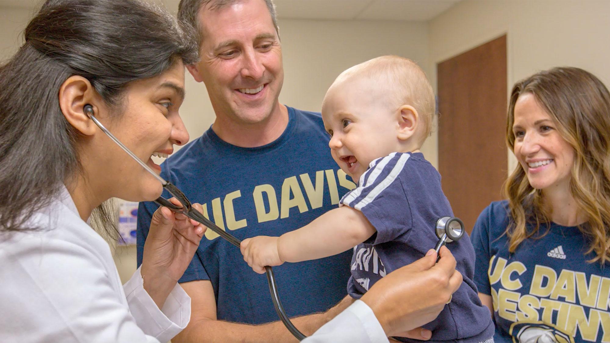 Two parents dressed in UC Davis Aggie gear present their baby to a doctor at a checkup