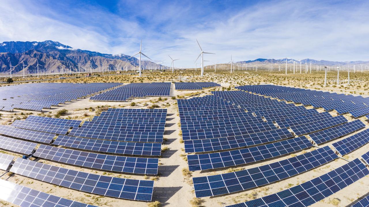 large area in desert covered by solar panels with windmills and mountains in background