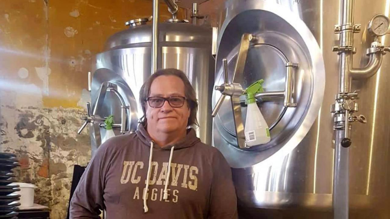 Glen Patrick Fox poses in UC Davis Aggies hoodie, amid brewing equipment.