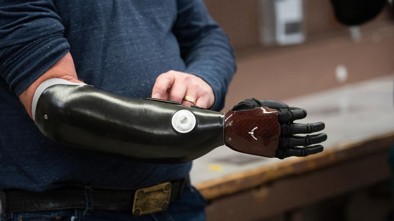 A black plastic prosthetic arm. The torso of the user, wearing a dark blue sweatshirt, is visible. 