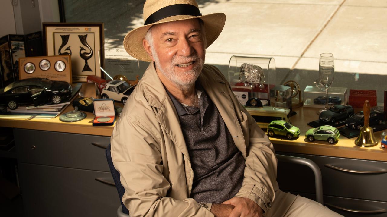 Inside an office sits a man in a straw fedora and beige jacket, smiling at the camera from a swivel chair in front of his desk, which has several different toy cars and other trinkets related to transportation. 