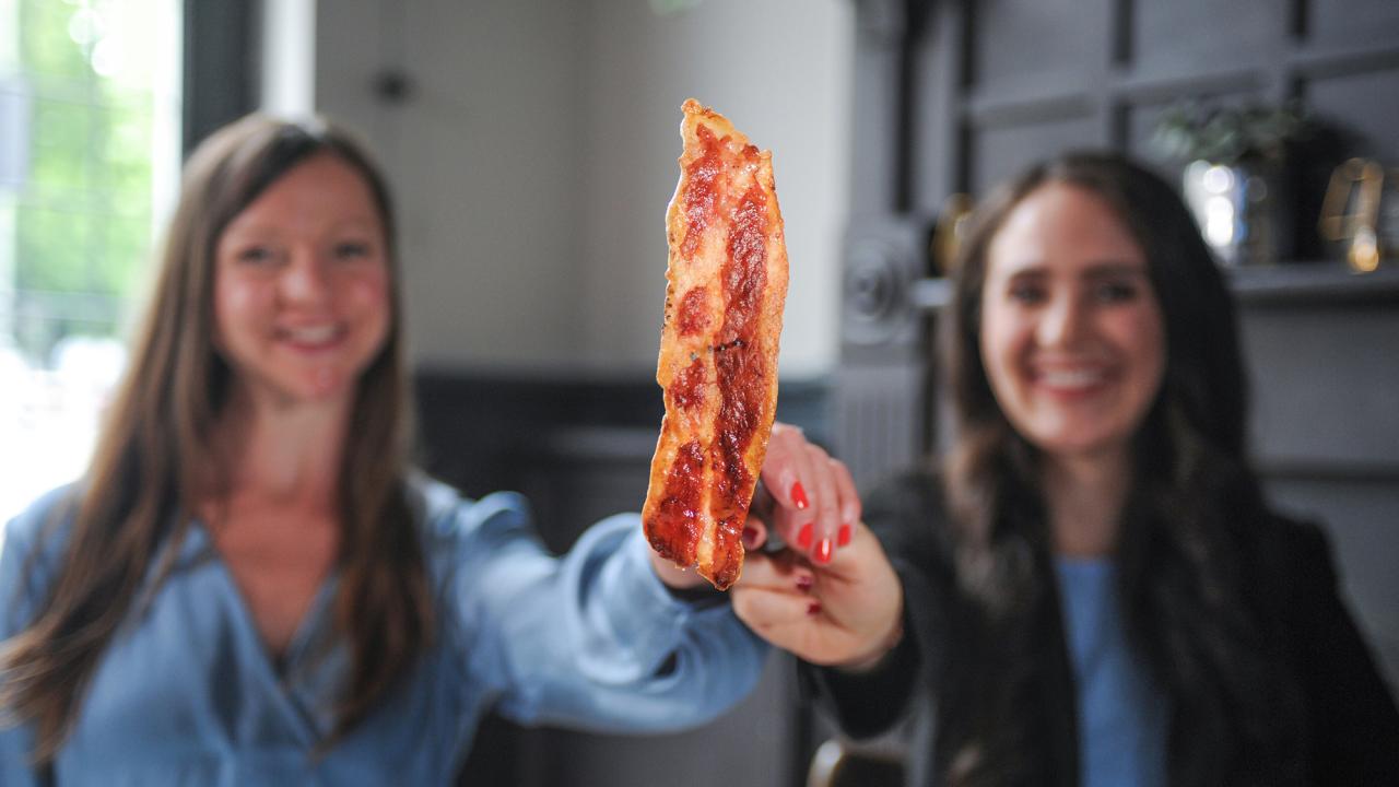 Two women hold up a cultivated meat item that looks like bacon