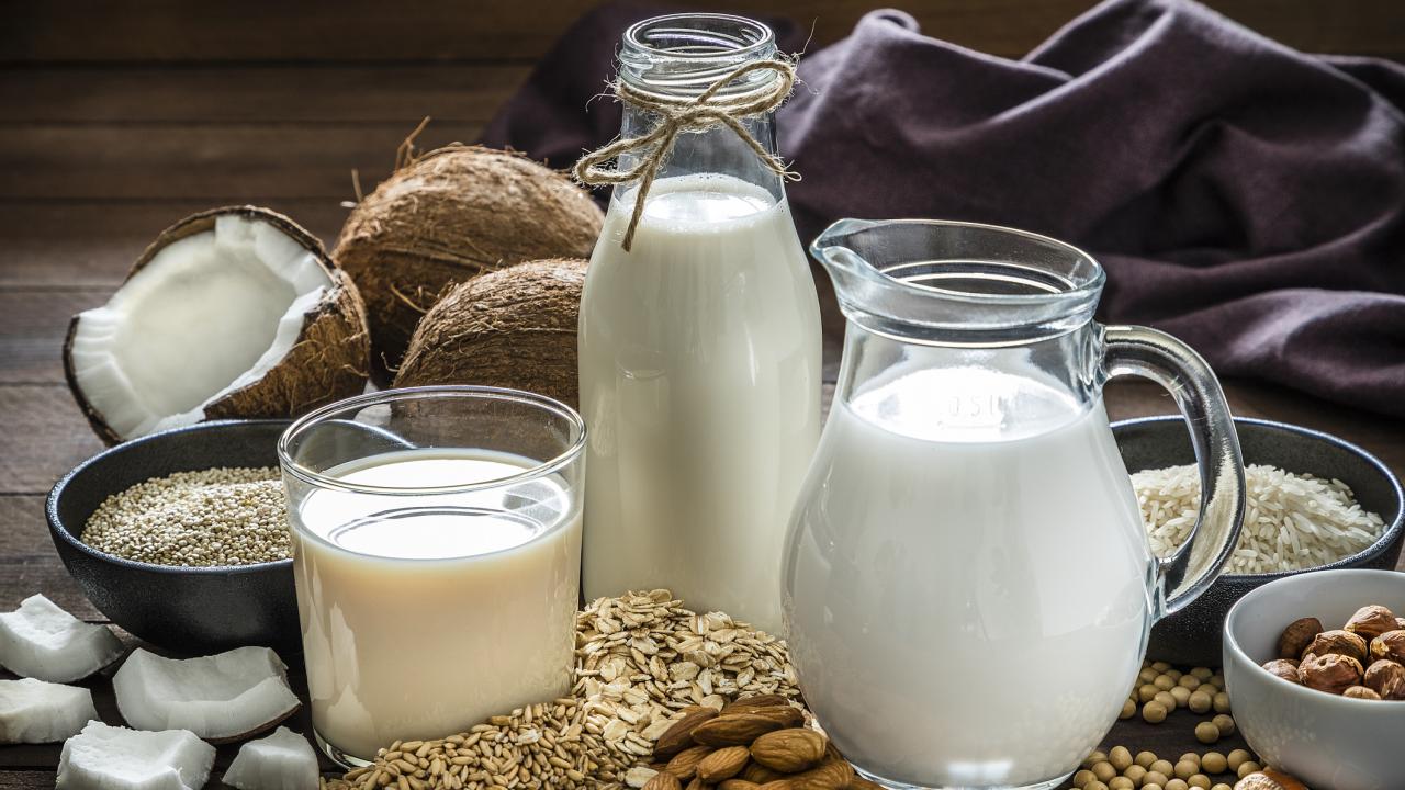 A glass, a bottle and a jug of milk surrounded by tree nuts, seeds, grains and coconuts. 
