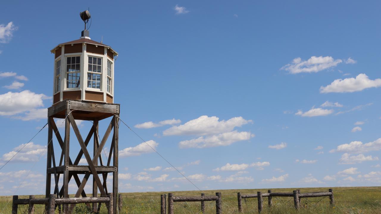 The guard tower at Amache National Historic Site, a Japanese Internment Camp in Colorado, rises high above the green grass around it on a bright, sunny day.